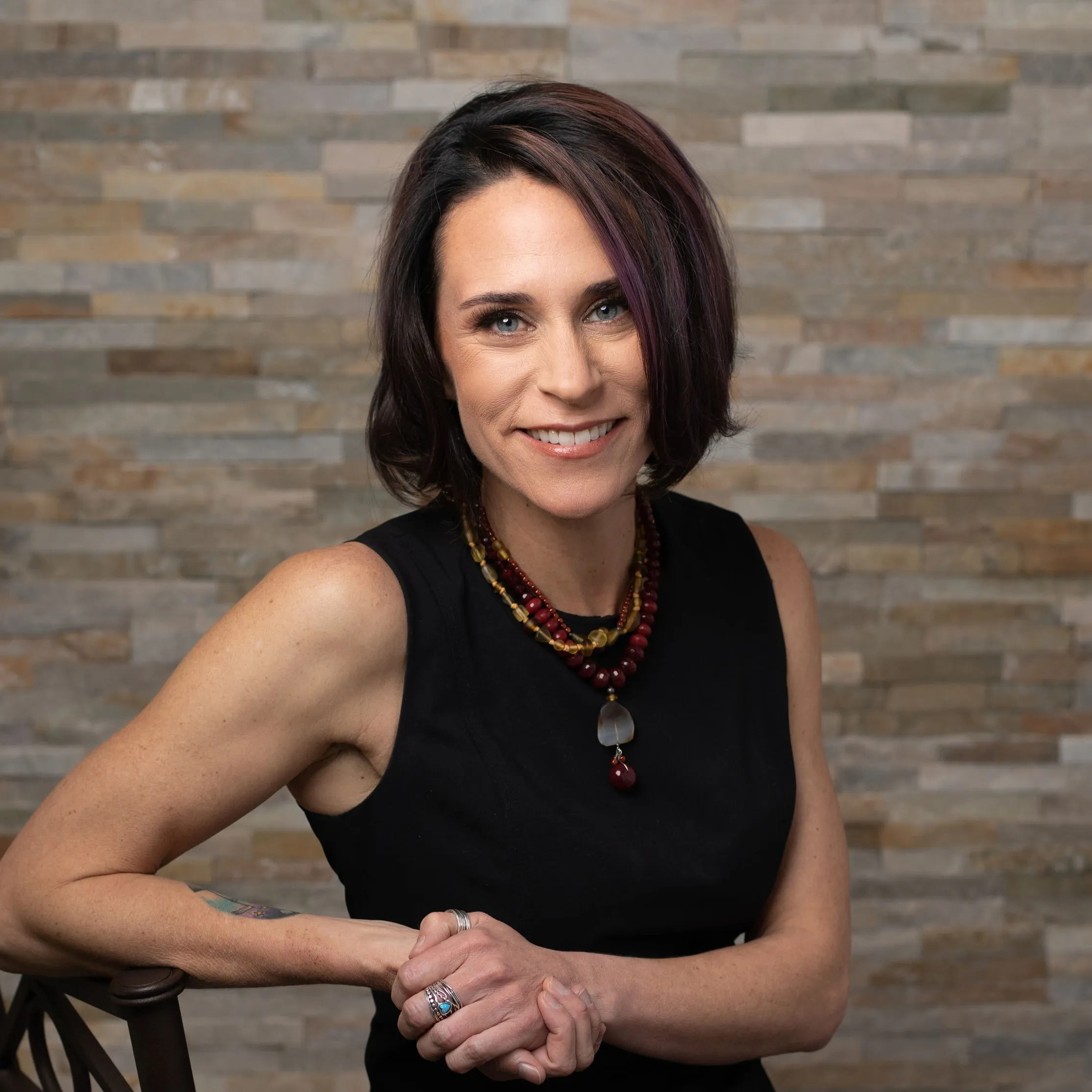 Smiling woman with short dark hair wearing a sleeveless black top and layered beaded necklaces, posing in front of a stone wall background.